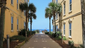 A paved pathway flanked by yellow buildings with palm trees, leading towards a distant view of the sea. - Adagio D405 Private Top 4th Floor Corner!! Premium Pool Front!+ Ice Maker! (Santa Rosa Beach)