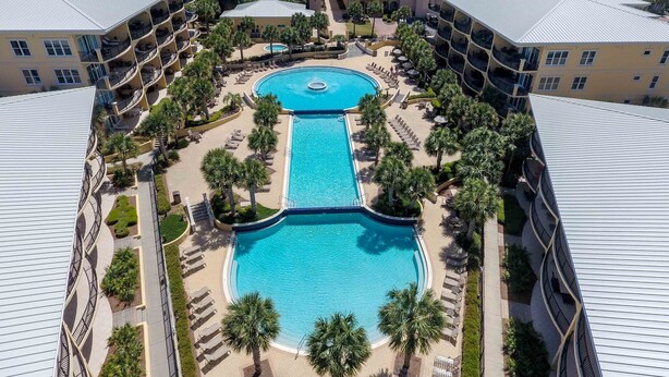 A resort with a central swimming pool surrounded by palm trees and lounge chairs, multiple hotel buildings with balconies, and a view of the ocean. - Adagio D405 Private Top 4th Floor Corner!! Premium Pool Front!+ Ice Maker! (Santa Rosa Beach)
