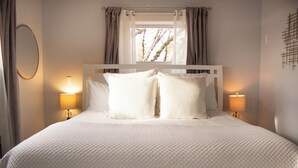 A neatly made bed with white bedding and pillows, two bedside lamps, a window with curtains, and a wall-mounted mirror.