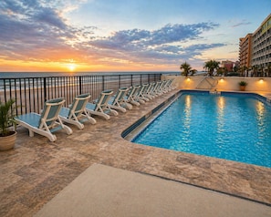 Área da piscina com espreguiçadeiras e vista para o pôr do sol no oceano.