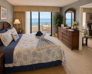 A bedroom with a large bed, a dresser, a mirror, a nightstand, a lamp, and a view of the ocean.