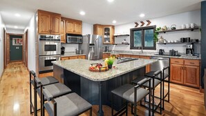 A modern kitchen with wooden cabinets, stainless steel appliances, a granite island with bar stools, and a breakfast bar. - Big Pine Alpine | Pickleball, GameRoom, HotTub, EpicViews, Gym & More (Laurelville)
