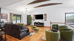 A spacious living room with a dark leather sofa, green armchairs, and a wooden floor. A flat-screen TV is mounted on the wall, and there is a staircase leading to an upper level. - Big Pine Alpine | Pickleball, GameRoom, HotTub, EpicViews, Gym & More (Laurelville)