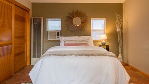 Une chambre à coucher comprenant un lit, un miroir fixé au mur, deux fenêtres équipées de stores, un radiateur et un vase décoratif.