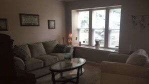 A living room with a beige sofa, a white armchair, a glass coffee table, and a window with blinds. - Charming home in Mountain Shadows. (Colorado Springs)