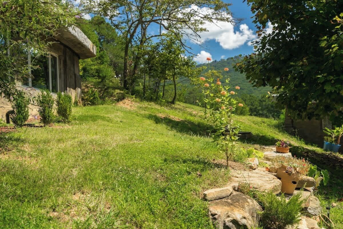 A rustic stone building with a wooden structure, surrounded by lush greenery and a hillside.