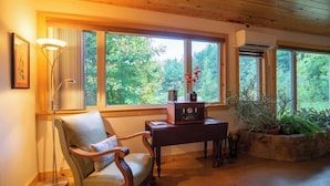 Une pièce chaleureuse avec un plafond en bois, une grande fenêtre donnant sur les arbres, un fauteuil, une table avec une radio et un lampadaire.