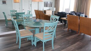 Un salon moderne avec une table à manger au plateau en verre, entourée de chaises turquoise, d’un canapé et d’un téléviseur fixé au mur.
