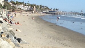 Ein Strand mit felsigem Ufer, Menschen, die sich sonnen, und ein Steg im Hintergrund.