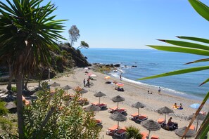 Ein Strand mit Reetdachsonnenstühlen und -schirmen, einer felsigen Küstenlinie und klarem, blauem Wasser.