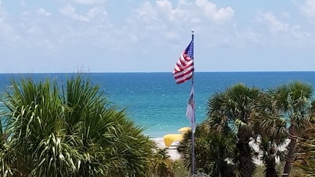 A flagpole with an American flag, a beach, and palm trees. - Gulf views! 2 Bedroom vintage condo - Third/Top Floor. FN #205 (St Pete Beach)
