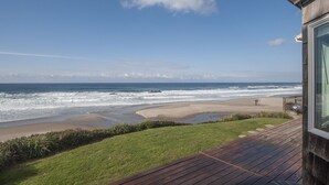 A beach view from a deck with a wooden railing, a grassy lawn, and a clear blue sky.