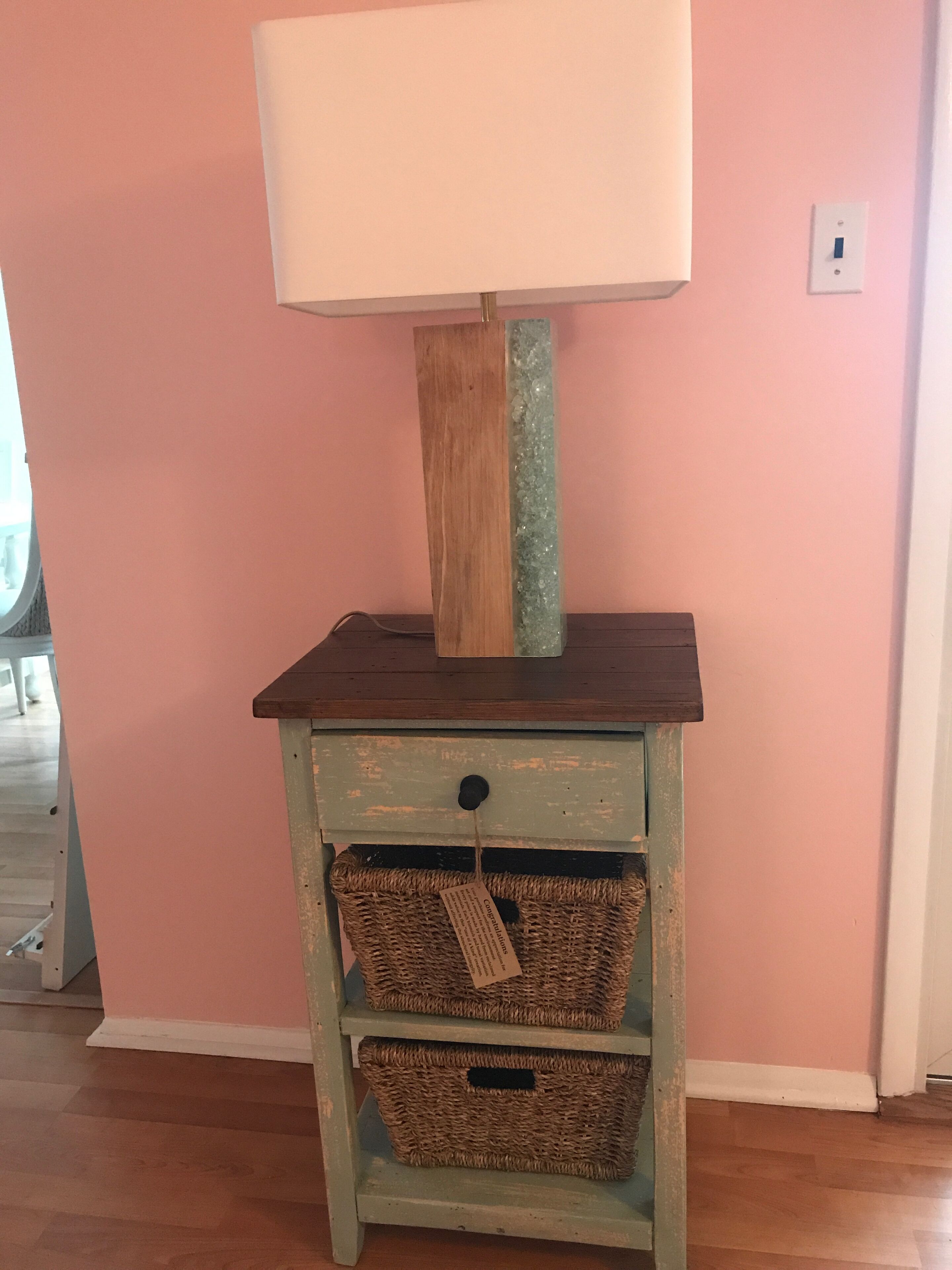 A wooden nightstand with a lamp, two wicker baskets, and a pink wall.