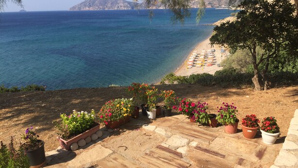 Un pati amb paviment de pedra i plantes en testos, amb vista a una platja d'ombrel·les i un paisatge de muntanyes al fons.