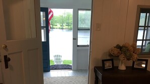 A room with a patterned rug, a wooden table with a vase of flowers, and a door leading to a balcony with a view of a body of water and trees. - Charming lake front cabin on Highland Lake (Andover)
