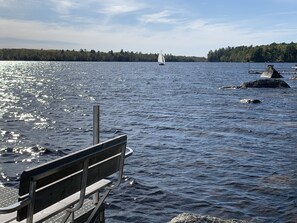 A wooden dock extending into a lake with a sailboat in the distance and a forested shoreline. - Cozy Haven: A Lakeside Cottage Near Acadia (Orland)