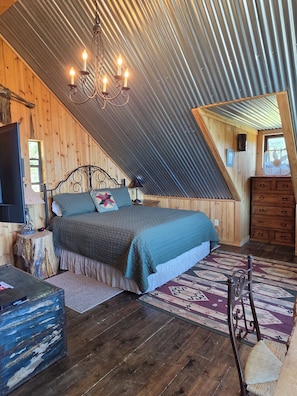 A bedroom with a bed, a chandelier, a dresser, a television, and a window.