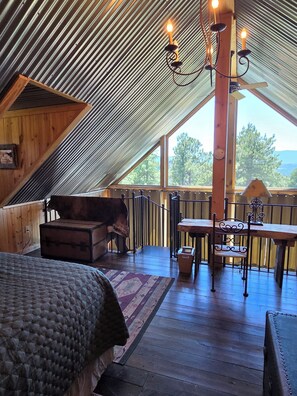 A cozy attic bedroom with a wooden bed, a small chest, a wooden table with a chair, and a view of trees through large windows. - Colorado Dream Vacation!5 min to Lake House!Next to nothing,close to everything! (Evergreen)