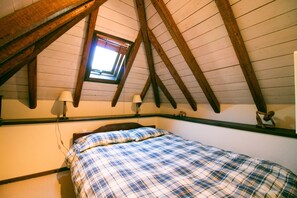 Une chambre avec un plafond en pente, des poutres en bois, un puits de lumière, un lit recouvert d’une couverture à carreaux, deux lampes de chevet et une petite étagère ornée d’un objet décoratif.
