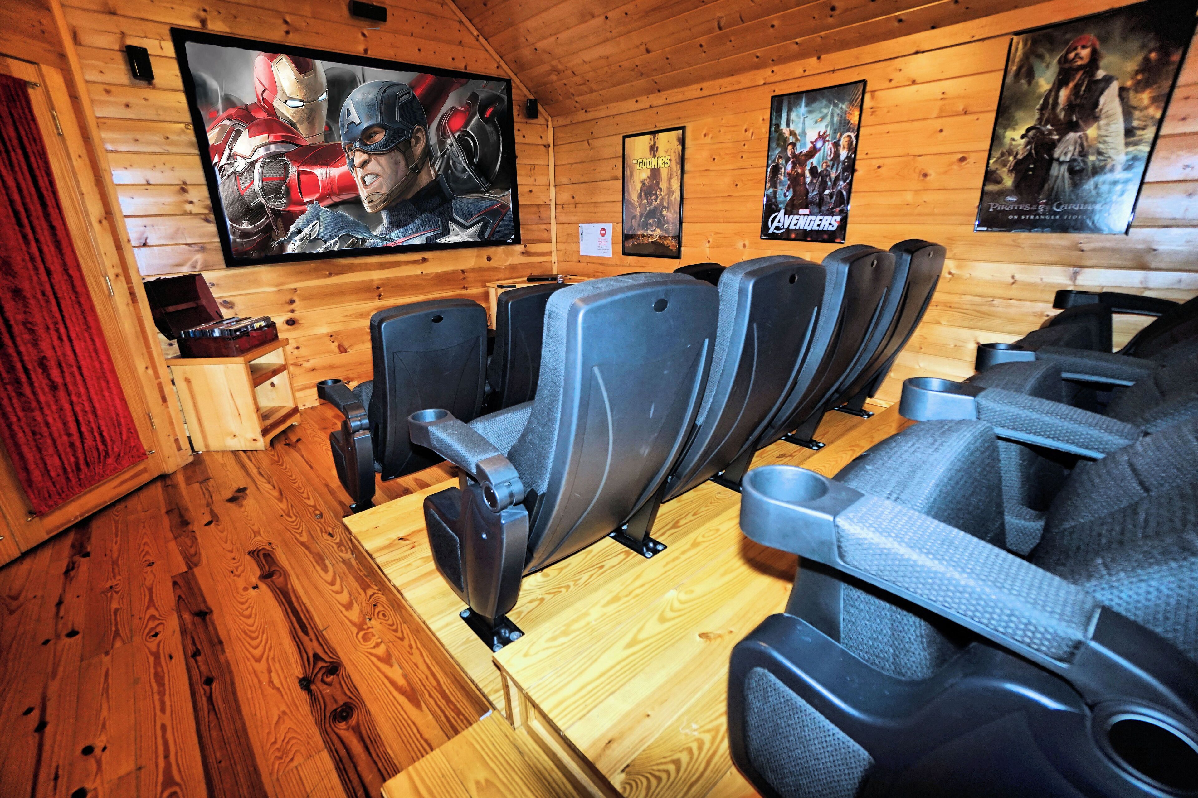 Una sala con paneles de madera, un televisor de pantalla plana grande, varios filas de asientos grises estilo teatro y carteles de películas en las paredes.