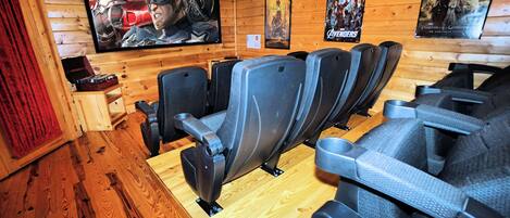 Una sala con paneles de madera, un televisor de pantalla plana grande, varios filas de asientos grises estilo teatro y carteles de películas en las paredes.