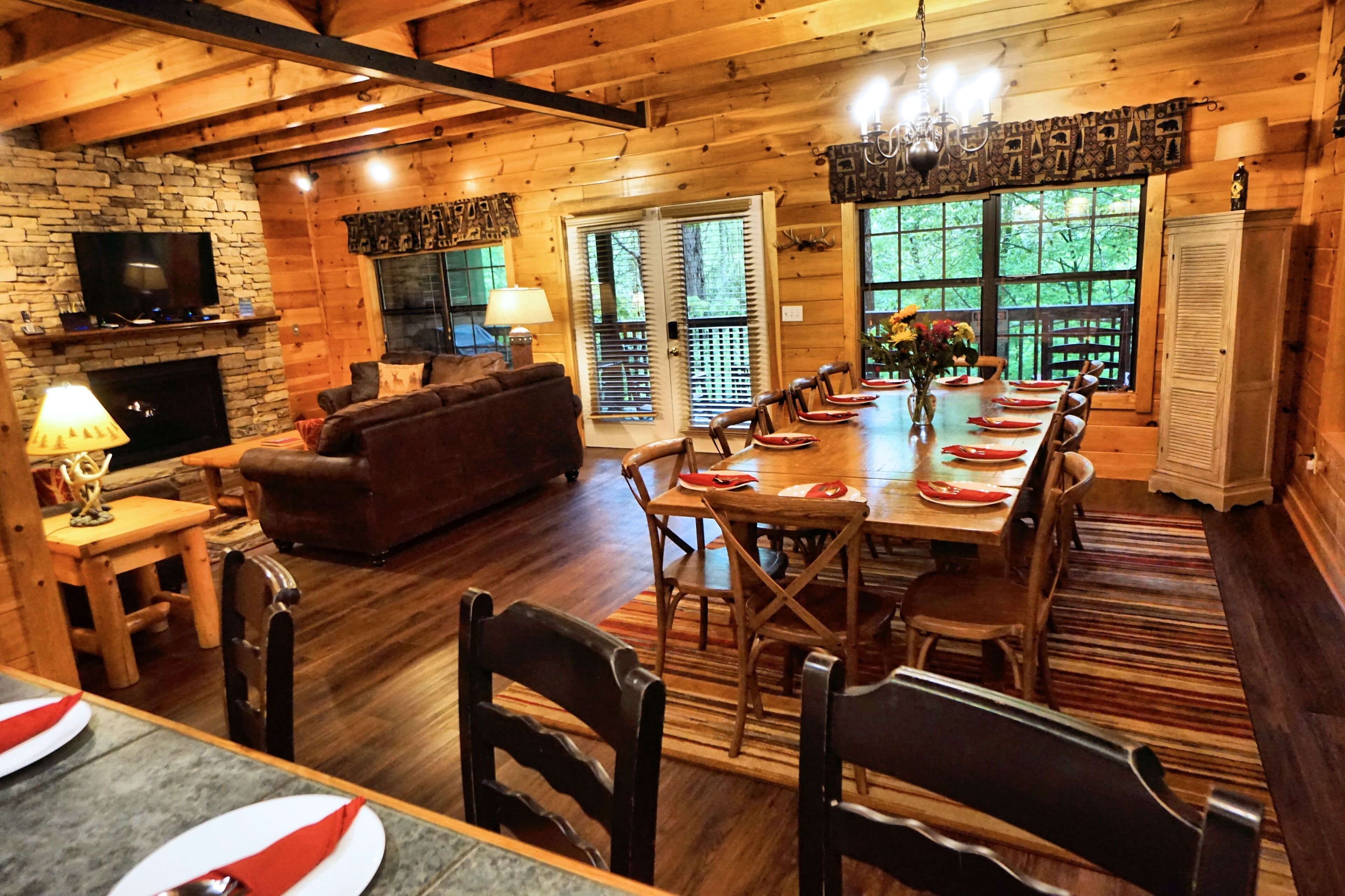 A wooden cabin interior with a dining area, fireplace, and large windows.