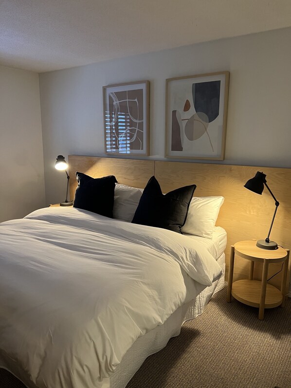 A neatly made bed with white linens and black pillows, a round wooden nightstand with a lamp, and two abstract art pieces on the wall.