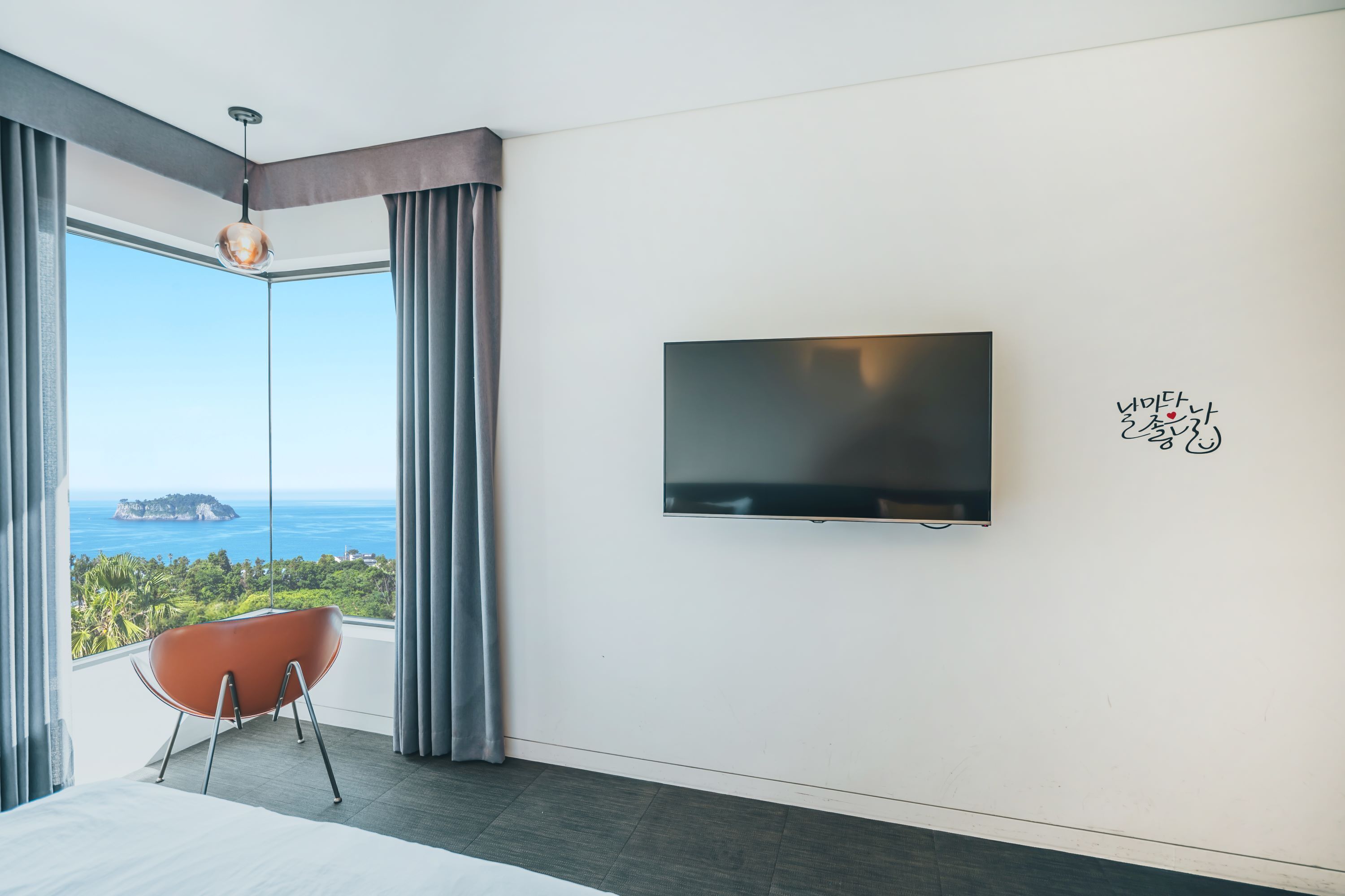 Habitación de hotel moderna con un ventanal que ofrece vistas al mar y a una isla distante. Un televisor de pantalla plana está montado en la pared, y una silla roja está colocada cerca de la ventana.