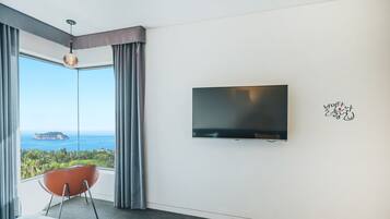 Habitación de hotel moderna con un ventanal que ofrece vistas al mar y a una isla distante. Un televisor de pantalla plana está montado en la pared, y una silla roja está colocada cerca de la ventana.