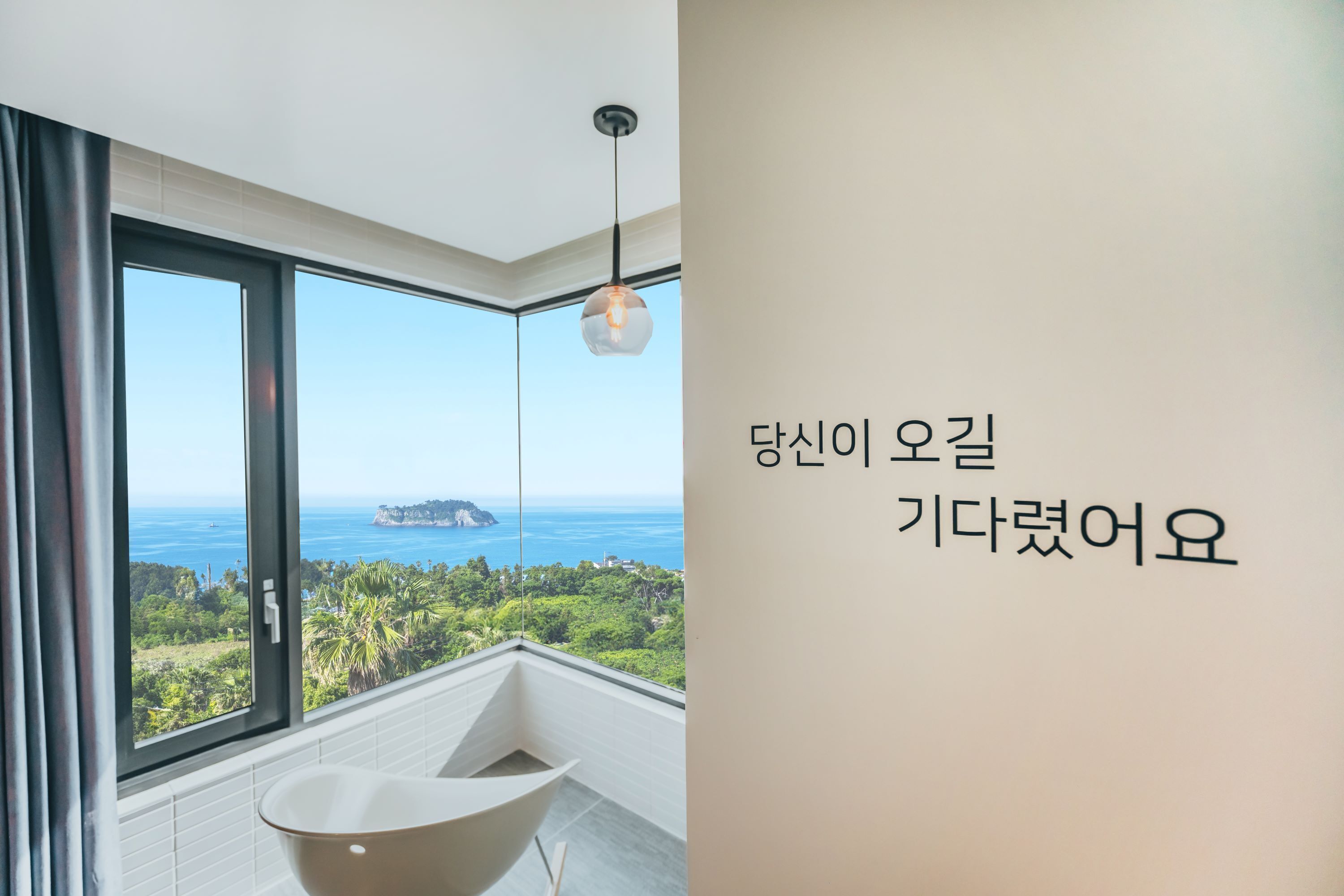A modern bathroom with a large window, a bathtub, and a hanging light fixture.