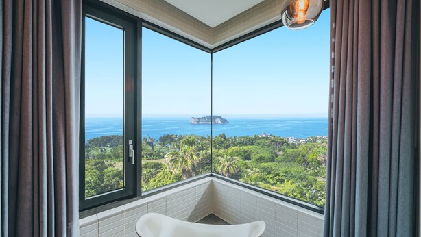 A modern bathroom with a freestanding bathtub, large windows offering a view of a coastal landscape, and a unique light fixture. - Jeju Bricks Hotel (Seogwipo)
