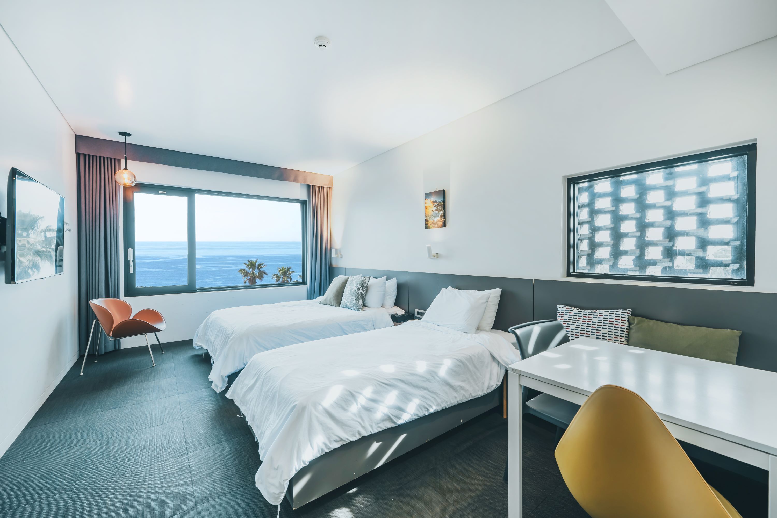 A modern hotel room with two beds, a dining table, and a view of the ocean.