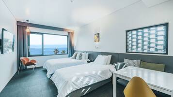 A modern hotel room with two beds, a dining table, and a view of the ocean.