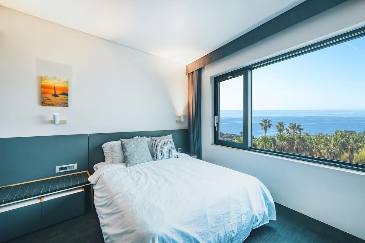 A modern hotel room with a large window offering a view of the ocean and palm trees.