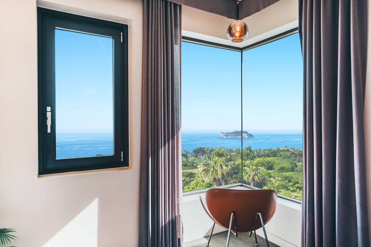 A room with a large window offering a view of the ocean and greenery.