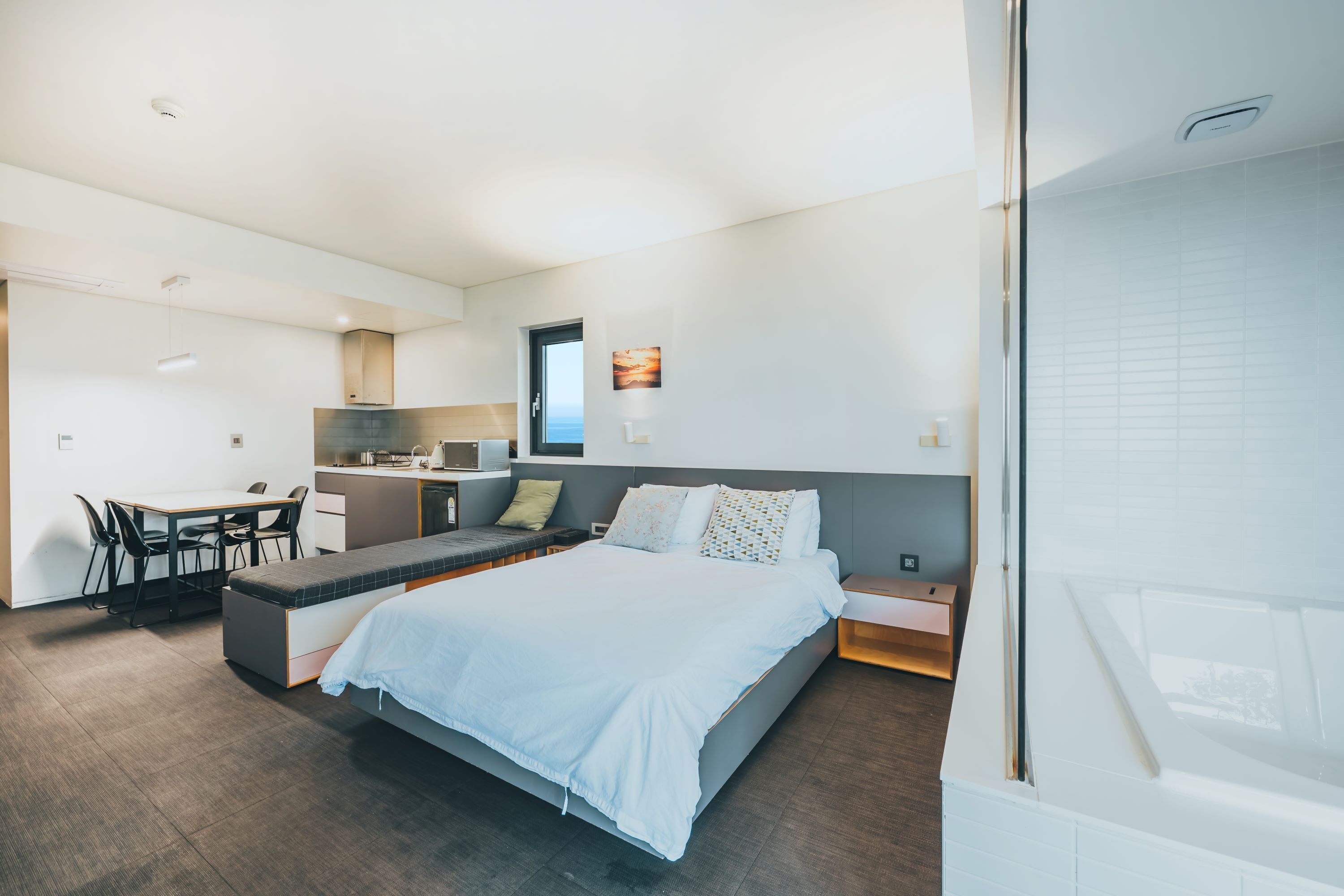 A modern bedroom with a bed, bedside tables, a desk, and a kitchenette.