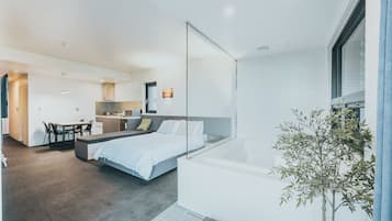 A modern hotel room with a large bed, a small desk, a kitchenette, and a view of the outside.