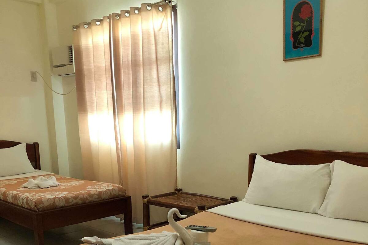 Habitación de hotel con una cama grande, una cama más pequeña, una mesita de noche de madera, aire acondicionado instalado en la pared y una ventana con cortinas.