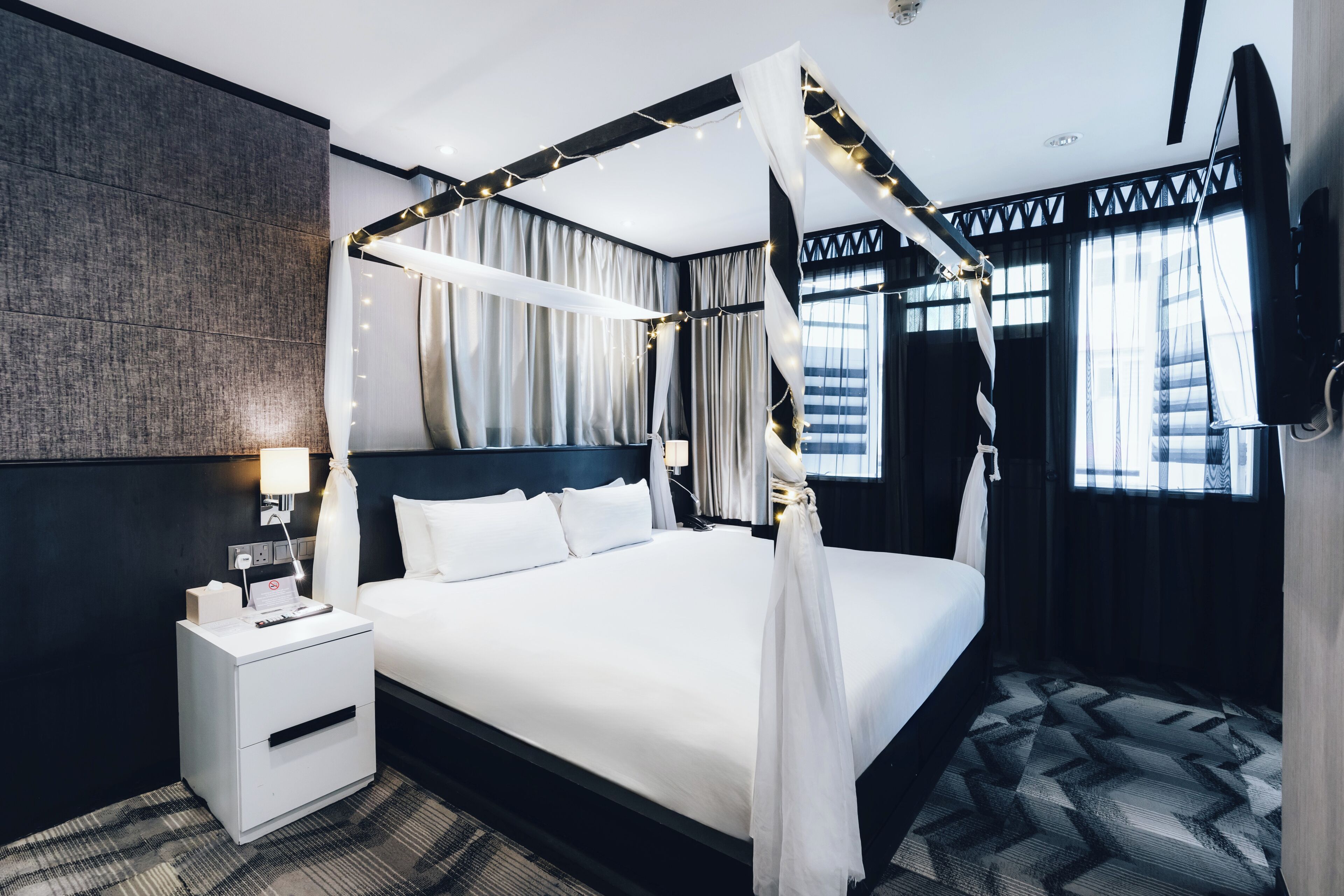 A modern hotel room with a four-poster bed, a nightstand with a lamp, and a large window with curtains.