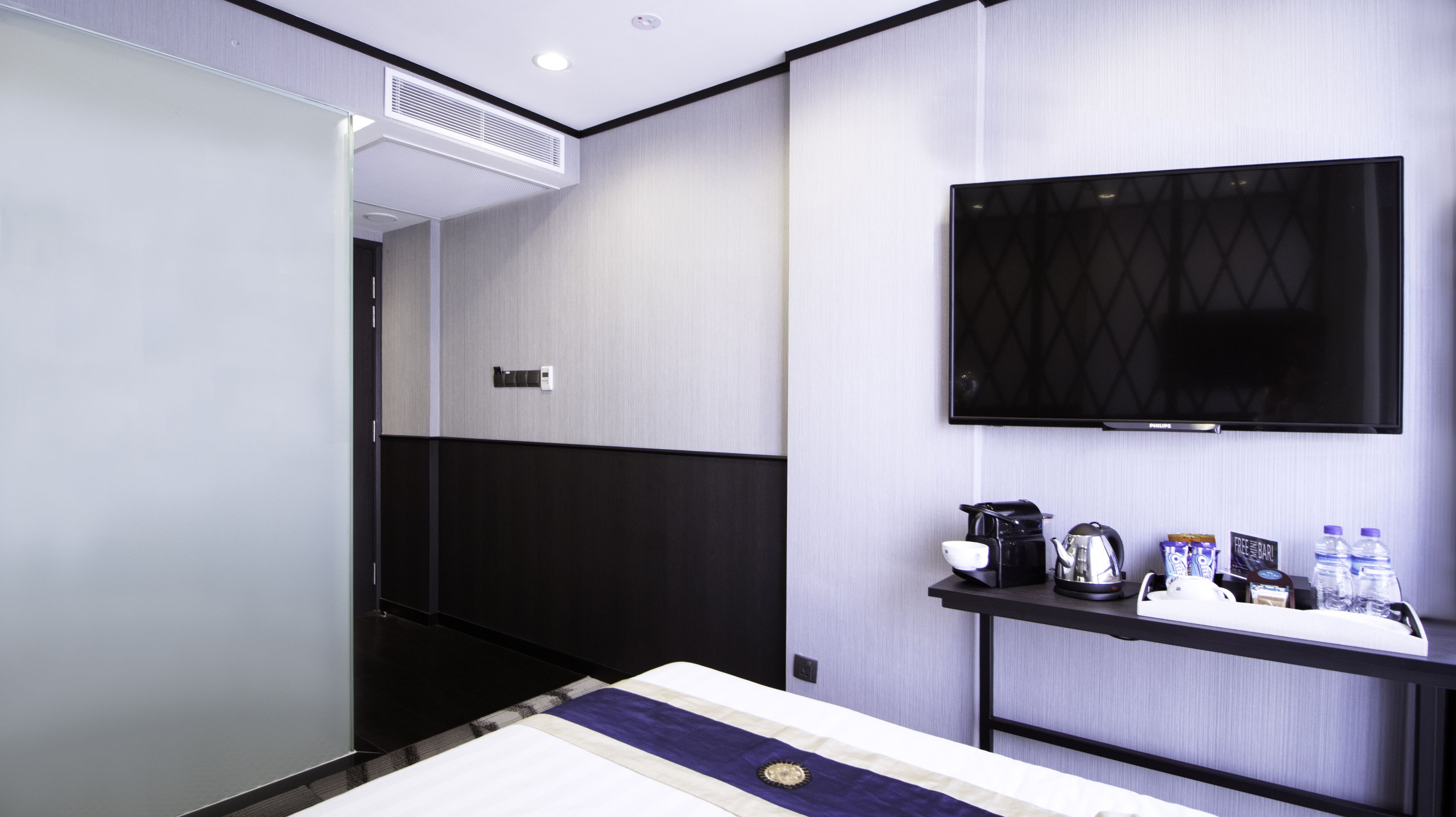 A hotel room with a bed, a TV mounted on the wall, a coffee maker, and a small table with water bottles and snacks.