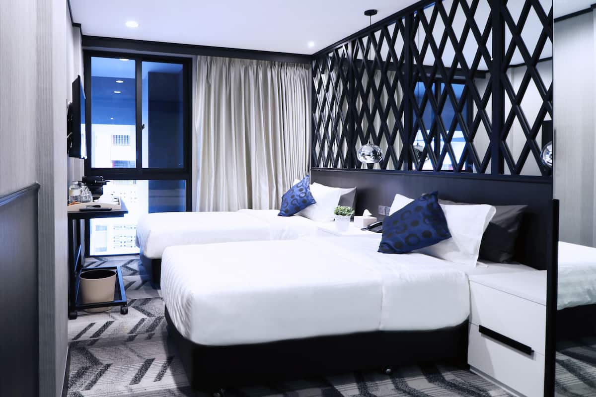 Une chambre d’hôtel avec deux lits, un bureau avec un ordinateur, une petite plante et une vue sur les bâtiments par la fenêtre.