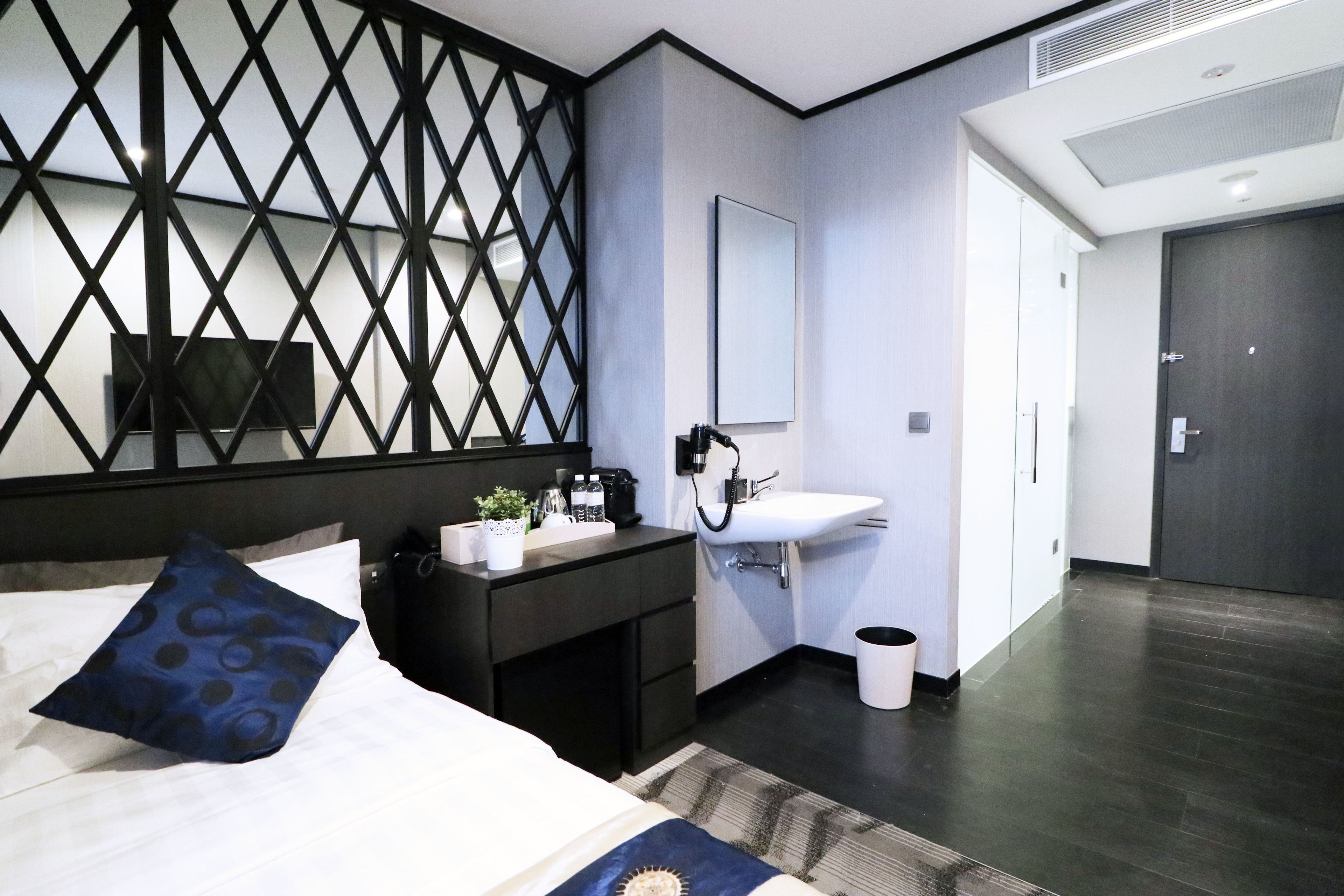 A modern hotel room with a bed, a desk with a sink, a mirror, and a dark wooden floor.