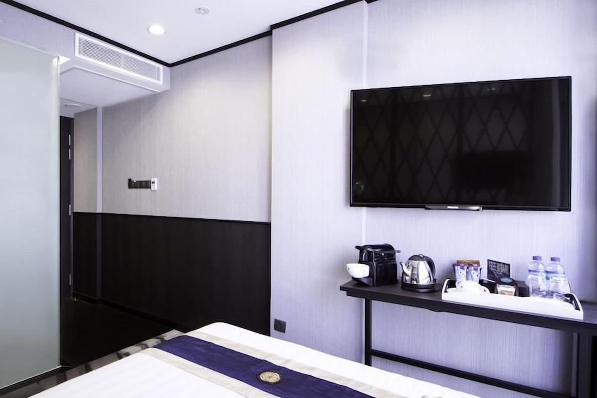 Une chambre d’hôtel comprenant un lit, un téléviseur à écran plat fixé au mur, une petite table avec une bouilloire et des bouteilles d’eau, et une porte donnant sur une autre pièce.