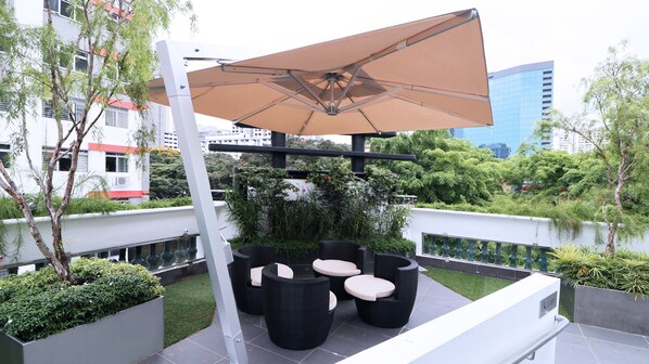 An outdoor seating area with a large umbrella, black wicker furniture, and a view of a cityscape with buildings and greenery.