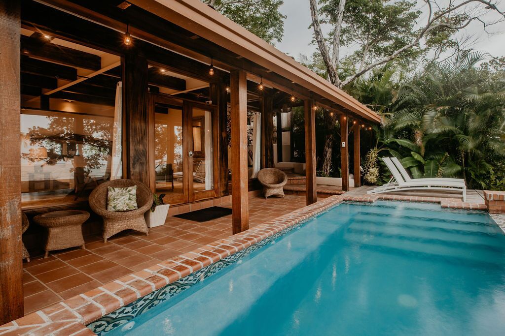 A covered outdoor area with a pool, wooden pergola, wicker furniture, and lush greenery.