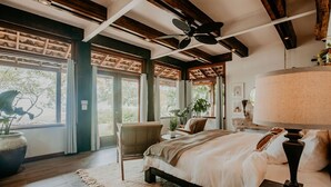 Un dormitorio con una cama grande, un ventilador de techo, una ventana con cortinas, una silla y una mesita de noche con lámpara.