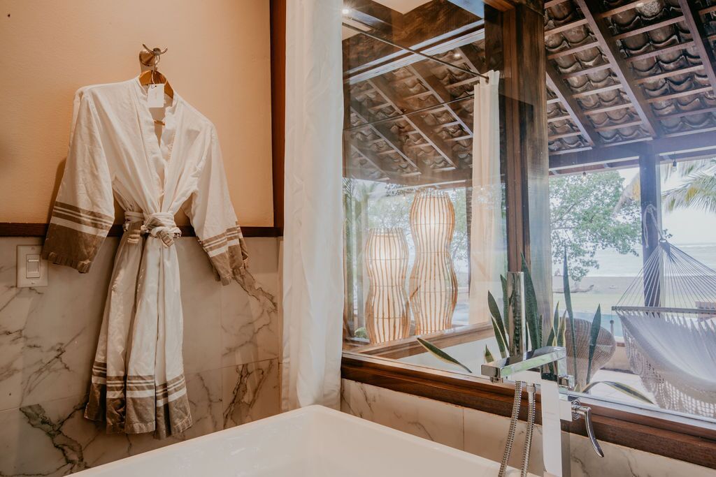 A bathroom with a marble wall, a white bathtub, and a window with a view of a hammock and plants outside.