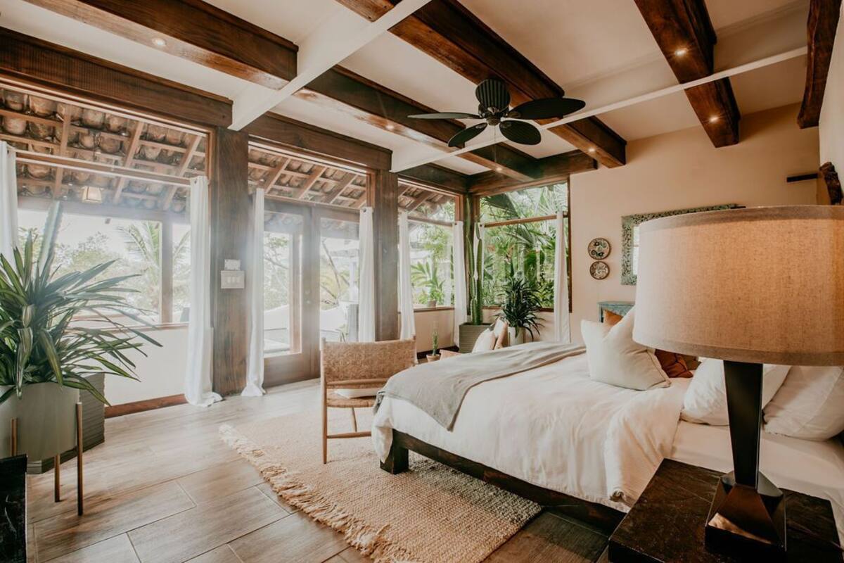 Un dormitorio amplio con una cama grande, un ventilador de techo y vistas al exterior a través de amplias ventanas.