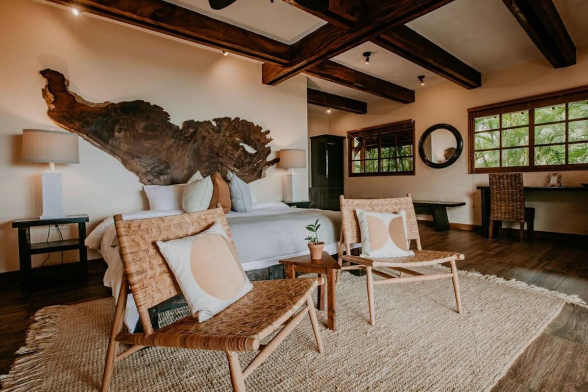 Un dormitorio con un gran cabecero de madera, una cama con ropa blanca, dos sillas de mimbre, una mesita y un ventanal con vistas a zonas verdes.