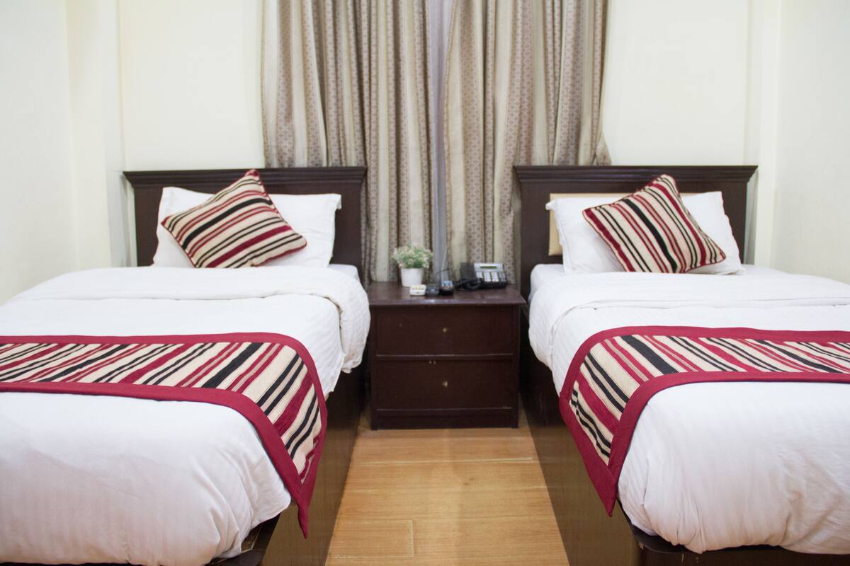 A hotel room with two single beds, each with a striped bedspread and matching pillows. There is a small bedside table with a lamp and a vase of flowers. The room has a wooden floor and a window with curtains.